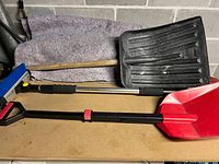 Three winter snow tools including one large standard black plastic snow shovel with wooden handle, one small adjustable red plastic shovel, and one snow brush with blue bristles on adjustable handle.