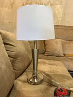 Side view of the silver bedside lamp with white lampshade placed on a brown couch surface.