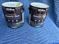 Two 1-gallon cans of BEHR Premium Plus Granite Grip Interior/Exterior paint primer in gray color sitting side by side on a blue padded surface.