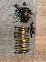 Top view of three sets of brass hardware including 12 curved brass handles, approximately 30 brass knobs, and a small set of antique handles, displayed on a gray mat with visible wood floor.