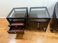 Two matching glass top side tables with visible felt-lined storage drawers and square dark wooden frames on hardwood floor.