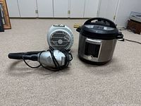 Three items visible on carpeted floor: Instant Pot Duo stainless steel pressure cooker with digital display, small white fan heater, and small handheld vacuum cleaner with hose attachment.