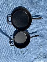 Two Lagostina cast iron frying pans side by side on a quilted blue blanket, one seasoned and lightly used, one new.