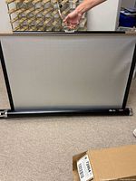 Photo showing the white retractable movie screen partially extended from its black metal casing with black border edges, held by a person's hand.