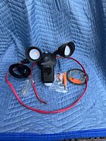 Overall view of the Ring Floodlight Cam Wired Plus with red wired power supply and included mounting hardware and accessories on a blue quilted fabric.