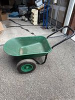 Green wheelbarrow showing full side view of basin, handles and wheels on pavement.
