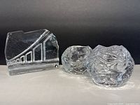 Photo showing two clear Kosta Boda Snowball glass candle holders and one Pukeberg Sweden geometric faceted glass paperweight against a dark background.