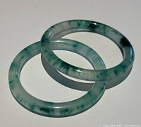 Two green and white translucent natural Asian jade fully round bangles, displayed overlapping on white surface, showing color variation and minor wear.