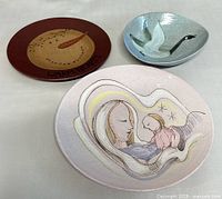 View showing three plates: a round wooden plate with snowman and text 'WARM HEARTS', a pale pink pottery plate with mother and child design, and a blue-gray pottery plate with a flying goose design.
