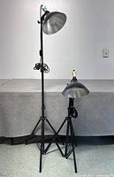Photo showing two black studio floor lamps, one fully extended and one collapsed, displaying adjustable tripod legs and dome-shaped silver shades.