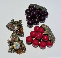 Two vintage Miriam Haskell scarf clips resembling grape clusters with large beads and brass metal leaf accents, alongside a pair of vintage clip-on earrings with rhinestones and decorative beads on a brass-tone filigree base.