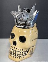Front view of beige ceramic skull mug with black detailing holding stainless steel cutlery including forks, spoons, and knives.