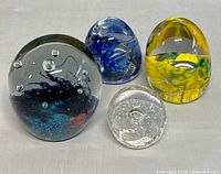 Four collectible glass art paperweights arranged together, showing various transparent designs with bubbles and swirls inside.
