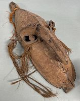 Front angle showing the mask’s long nose, eye sockets with wooden spheres, and fiber tassels hanging from sides.