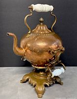 Front view of vintage copper teapot on brass stand with white porcelain handle on top and base.