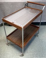 Front and side view of the teak and metal rolling tray cart with built-in electric heating element on top tray surface. Shows power cord and caster wheels.