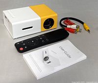 Mini portable LED projector with remote, AV cable, and user guide on gray surface.