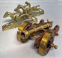 Photo showing two brass racehorse and rider figurines positioned behind the brass and wood decorative cannon.