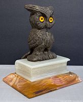 Front side showing metal owl with yellow eyes perched on a book-shaped white marble base atop a polished orange-pink marble slab.