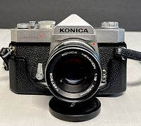 Front view of KONICA AUTOREFLEX T film camera with attached 52mm f/1.8 HEXANON lens and lens cap on a white table surface.