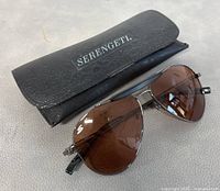 Serengeti aviator style sunglasses positioned in front of the black leatherette case with logo on flap visible.