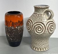 Photo showing two West German vases side-by-side, one beige with swirl patterns and handle, the other orange lava glaze cylindrical vase.