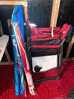 View showing two folded red camping chairs and two umbrellas in cases, one blue and one red and white in plastic wrap, all stacked together.