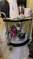 Full view front of round bar or coffee cart with black surfaces, chrome frame, and wheels, with various items placed on shelves for size perspective.