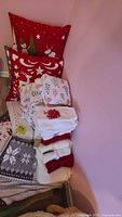 Two red Christmas decorative pillows with holiday patterns, folded sheets and linens in white with red and green Christmas tree and holly designs, plus folded towels in white, red, and gray.