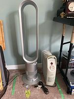Dyson bladeless tower fan in silver finish next to cream-colored Airworks oil heater with remote on floor