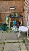 Wide view of assorted garden tools and accessories including hose, watering cans, gloves, long handled tools, and a plastic stool.
