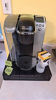 Full view of the Keurig K70 coffee maker with water reservoir on the left, control panel on the right, accompanied by a K-Cup holder sitting below and a small white cup.