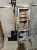 Aluminum ladder with three steps leaning against wall, next to three shovels (one metal scoop shovel and two plastic snow shovels) standing upright.