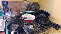 Multiple cookware pieces stacked including pots, a pizza stone, frying pans, and kitchen racks on a stove.