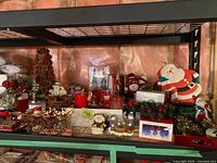 Shelf with assorted Christmas-themed decorations including floral arrangements, snow globes, figurines, candles, and Santa figurine.