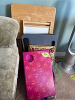 Stacked TV trays and lap desks resting next to a couch including wood, plastic, and decorative styles
