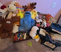 Photo shows a group of plush toys including Cookie Monster, brown moose, Snoopy, and yellow chicks, with a box of toy cars in front and some stacked books and toys on the right side.