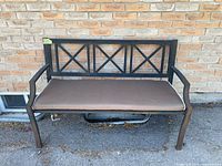 Full view of outdoor bench with black metal frame and brown removable seat cushion against brick wall background.