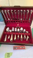 Full view of stainless steel flatware set inside red lined case. Utensils neatly arranged in slots with knives on upper lid, and forks, spoons, dessert forks, and demitasse spoons below.