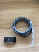 Rolled black leather belt in plastic wrap beside LV-logo buckle