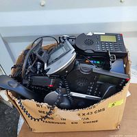 Photo shows a box filled with five black office desk phones with handsets, coiled cords, and digital screens.