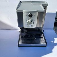 Front view of stainless steel Breville Cafe Roma espresso machine showing power and heating indicator lights, control dial, drip tray, and power cord wrapped on machine.