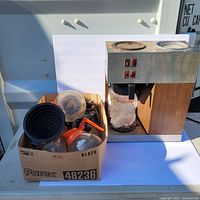 Photo of Bunn-O-Matic coffee maker with woodgrain and stainless steel design, two warming plates on top and control switches, sitting on a white table next to a box filled with various coffee pots and accessories.