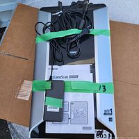 Top-down view of the Canon CanoScan 8800F scanner with cables and power adapter attached, secured with green tape on the closed lid. The user manual is visible under the tape.