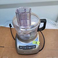 KitchenAid food processor in silver with clear 7-cup work bowl and black handle, showing full unit from top front angle.