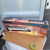 Photo of the Yamaha PSR-75 keyboard packed in its original box with protective wrap and some visible wear on the box.