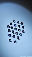 22 loose round calibrated cornflower blue sapphires arranged on a white surface with visible diamond cuts and vivid blue color under lighting.