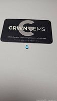 Photo of single small teardrop-shaped Swiss blue topaz gemstone placed below a CRWN Gems business card for scale and brand reference.