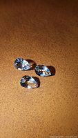 Three pear-shaped aquamarine gemstones lying on a brown surface, showing light blue color and facets.