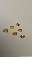 Photo showing six natural golden yellow sapphires arranged on a white background, five pear cuts and one round cut, showcasing their golden yellow hues and clarity.